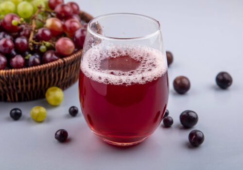 side view of black grape juice in glass and basket of grapes with grape berries on gray background